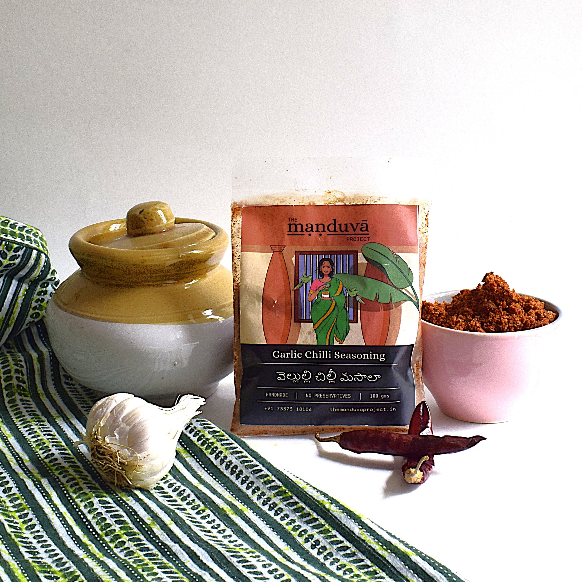 Packaging of Manduva Garlic Chilli Podi Sprinkle with a bowl of the spice mix and whole garlic cloves in the foreground.