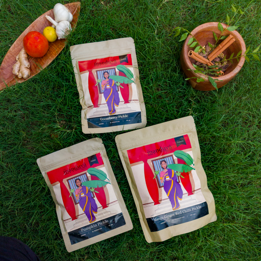 Three packets of Manduva Seasonal Favourites pickles, featuring Mango Ginger, Gooseberry, and Pumpkin, placed on a grassy surface with a plant and some food items in the background.